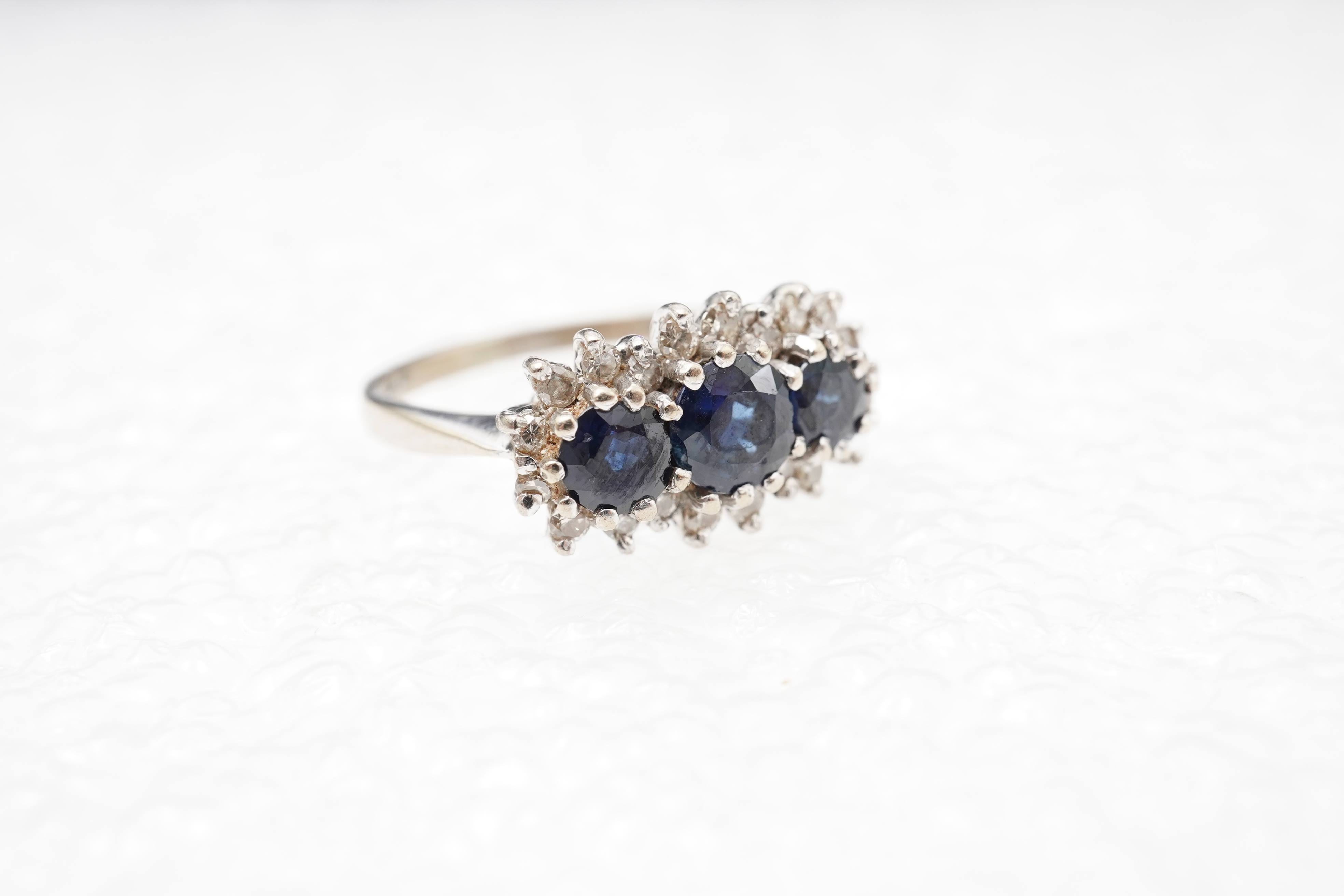 A sapphire and diamond ring, set with three circular-cut and oval sapphires, within a border of brilliant-cut diamonds totalling approximately 0.40 carats, mounted in 18ct white gold, size M1/2, British hallmarks, gross